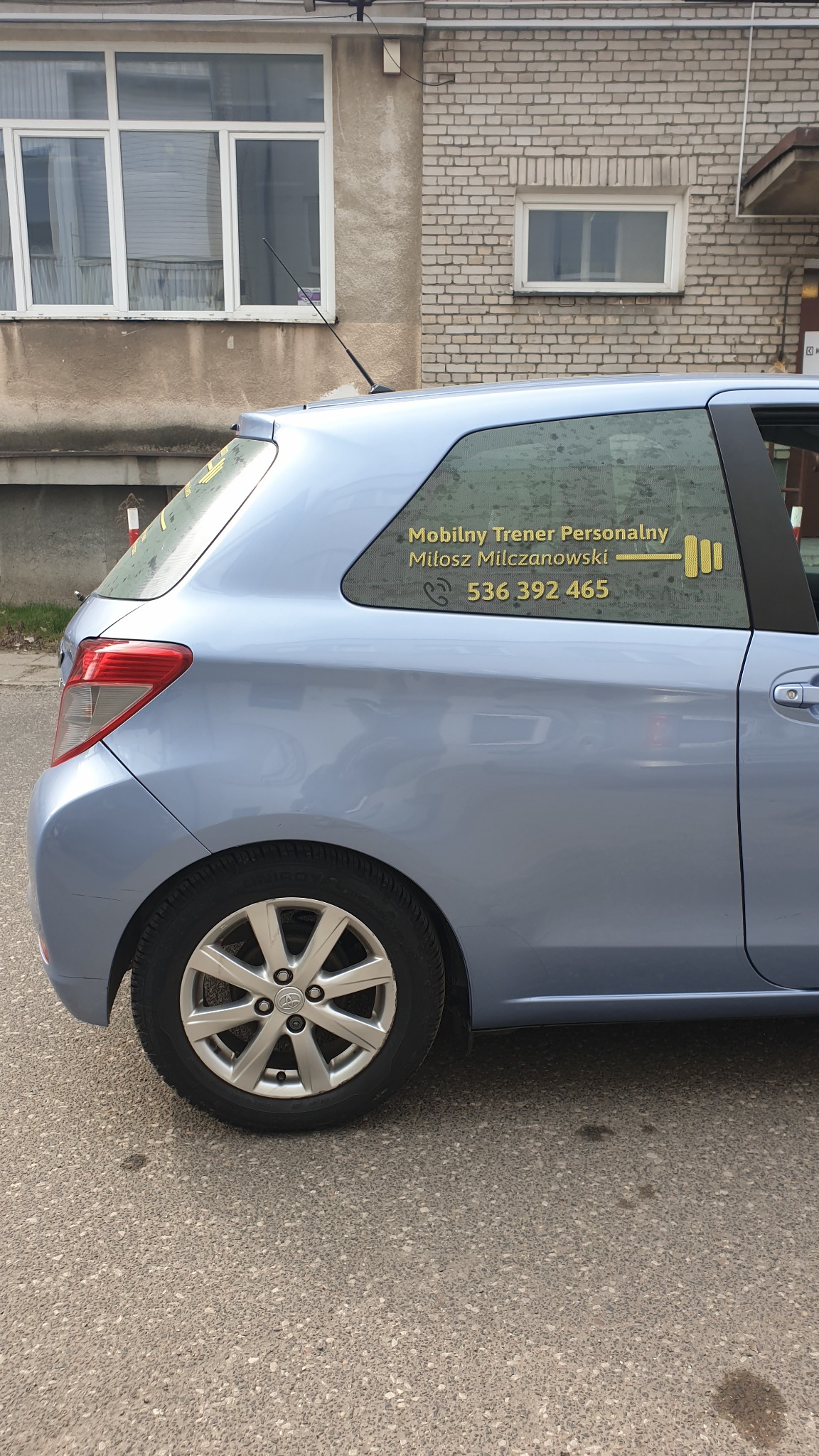 Niebieski samochód z folią OWV na tylnej szybie, prezentującą dane kontaktowe mobilnego trenera personalnego oraz symbol sztangi w kolorze złotym.