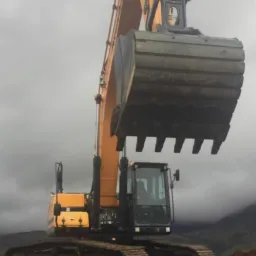 Żółto-czarna koparka hydrauliczna na tle pochmurnego nieba i odległych gór, stojąca na nierównym terenie z ciemną ziemią.