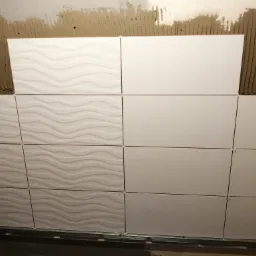 Układanie białych, prostokątnych płytek ściennych z wzorem falistym i gładkich, z użyciem listwy startowej i krzyżyków dystansowych, na ścianie z widocznym klejem.