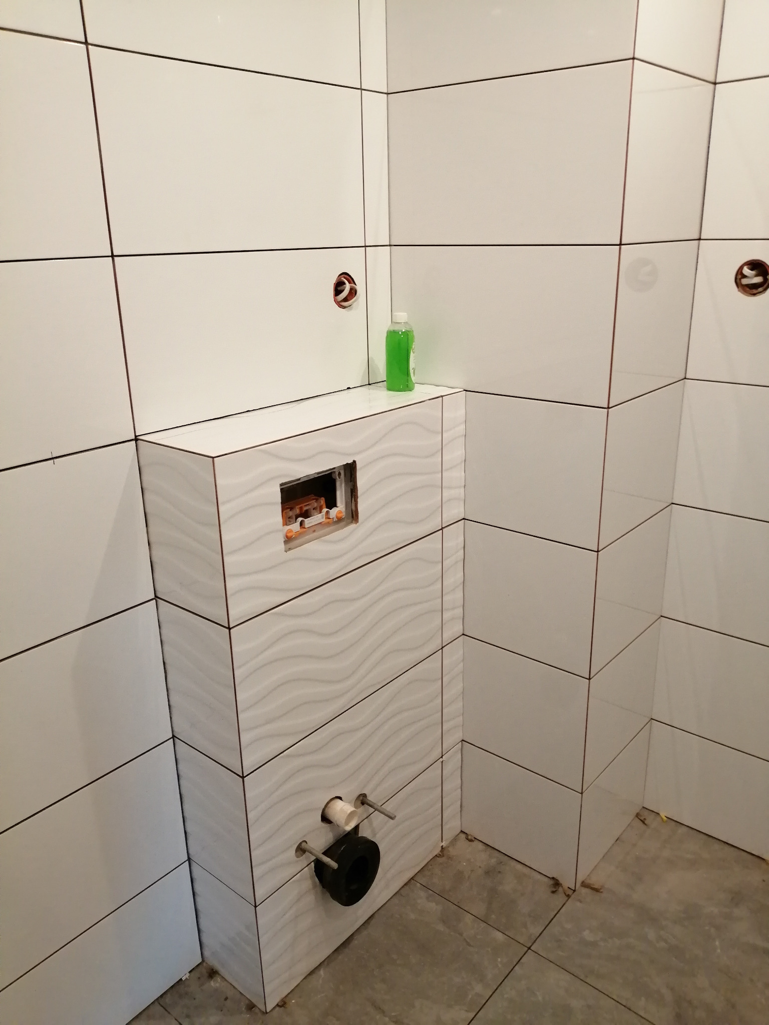 Białe, prostokątne płytki na ścianie i zabudowie stelaża podtynkowego WC z widocznymi elementami instalacji. Na górze zabudowy stoi butelka z zielonym płynem.