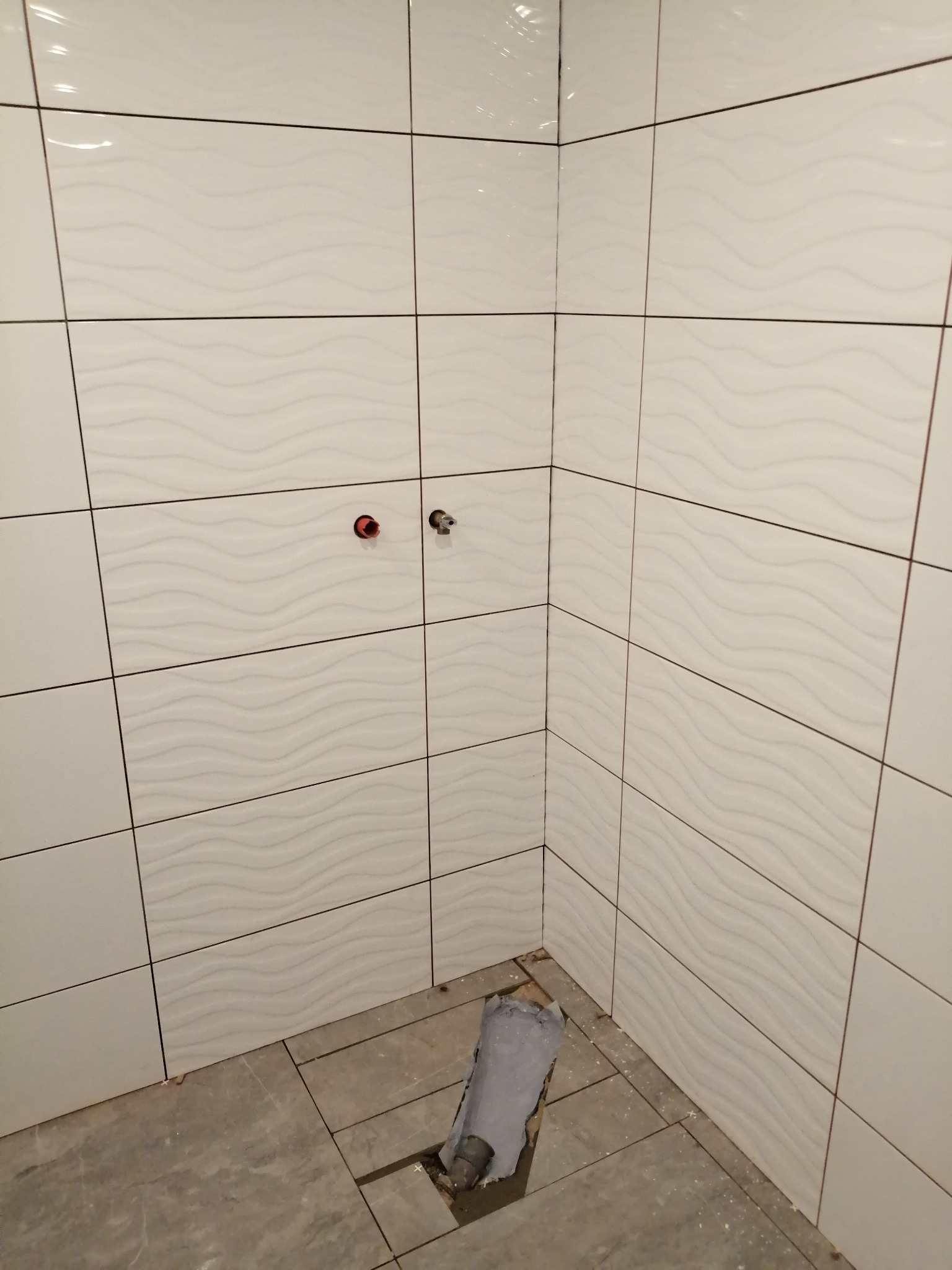 Ściana w trakcie wykańczania z białymi, prostokątnymi płytkami z delikatnym falistym wzorem, widoczne otwory na armaturę i częściowo ułożona podłoga z szarych płytek.