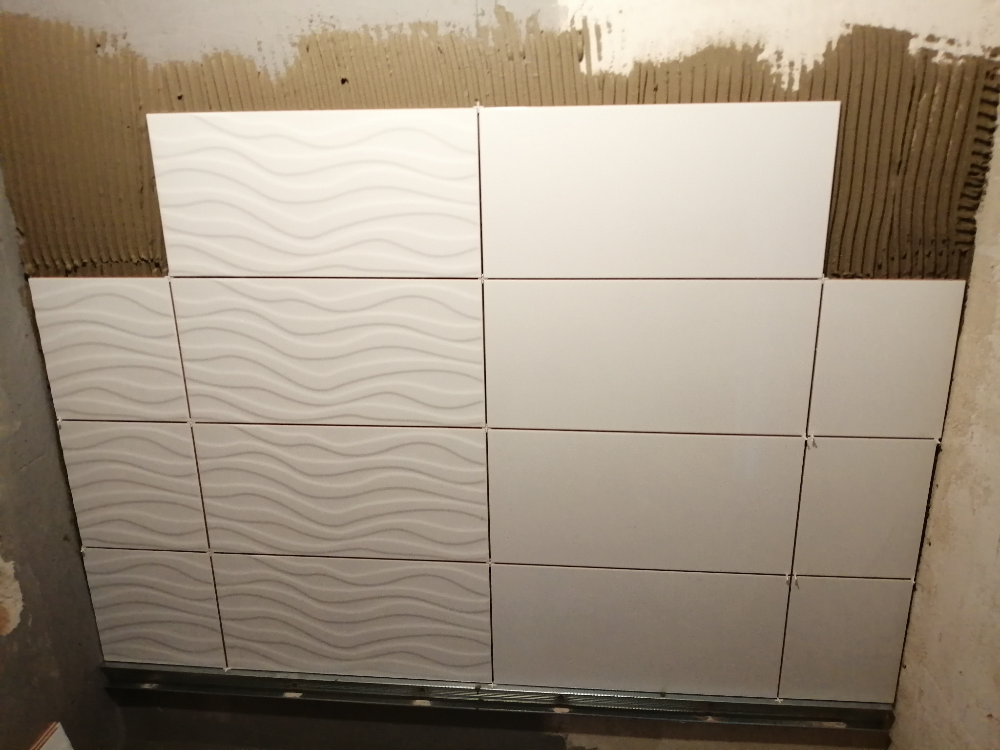 Układanie białych, prostokątnych płytek ściennych z wzorem falistym i gładkich, z użyciem listwy startowej i krzyżyków dystansowych, na ścianie z widocznym klejem.