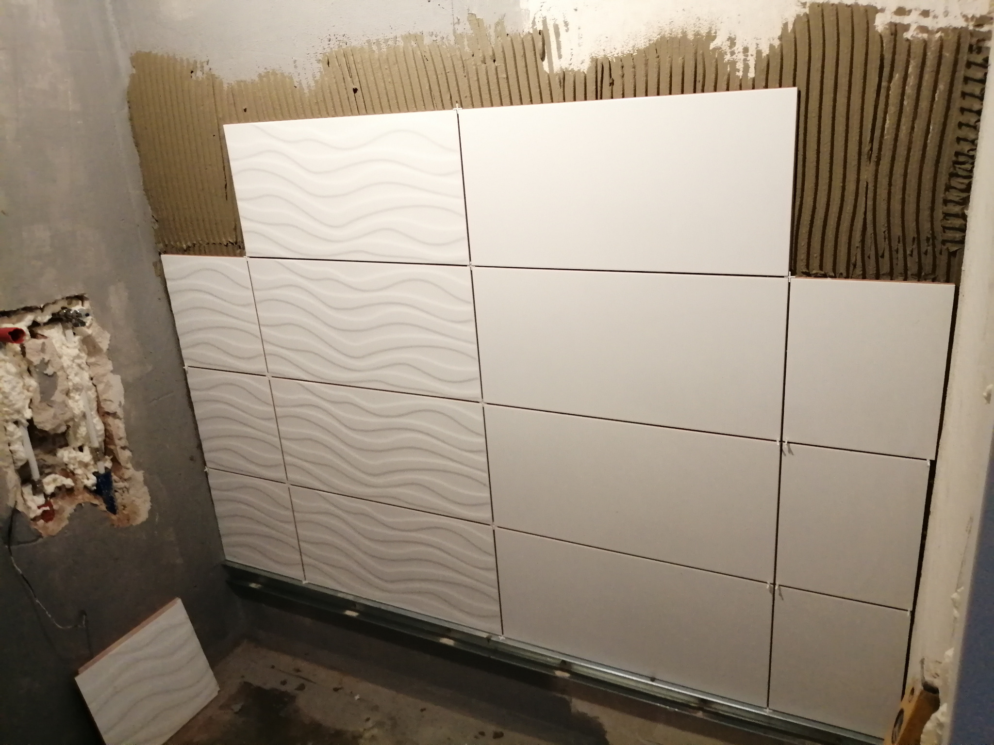 Początkowy etap układania białych płytek ściennych z delikatnym wzorem falistym, widoczne szyny montażowe, świeży klej na ścianie i niezabezpieczone rury w ścianie obok.