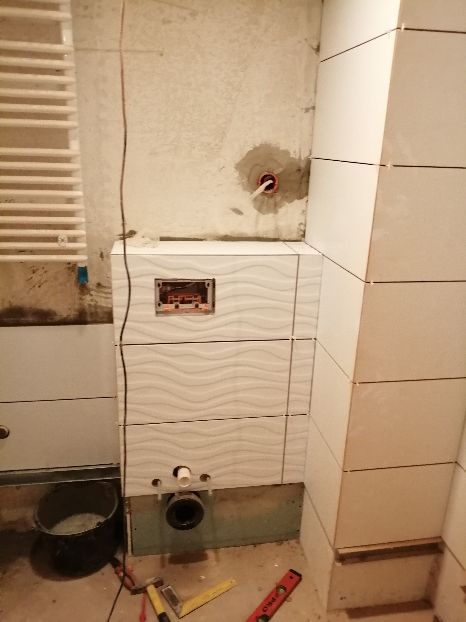 Montaż podtynkowego stelaża WC z białymi płytkami o falistym wzorze w trakcie prac wykończeniowych, widoczne narzędzia i wiadro z zaprawą.
