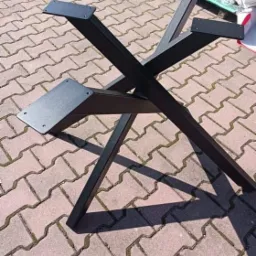 Metalowa podstawa stołu w kształcie litery X, pomalowana na czarno, wykonana w Alwerni, widoczna na tle kostki brukowej.
