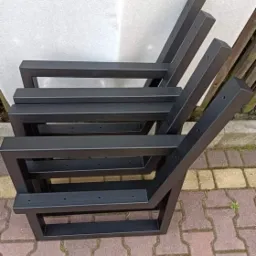Stos czarnych, spawanych, metalowych nóg do krzeseł z otworami na śruby, ustawionych na kostce brukowej.
