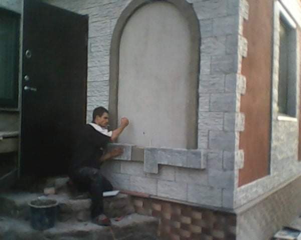 Mężczyzna w trakcie układania kamiennej elewacji wokół łukowatego okna na budynku. Widoczny fragment ściany z oknem, drzwi wejściowe i wiadro na schodach.