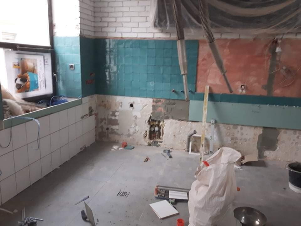 Wnętrze w trakcie układania płytek: białe płytki na ścianie pod oknem, turkusowe płytki na sąsiedniej ścianie, odsłonięte fragmenty ściany z instalacjami, narzędzia na podłodze, worek z materiałami...
