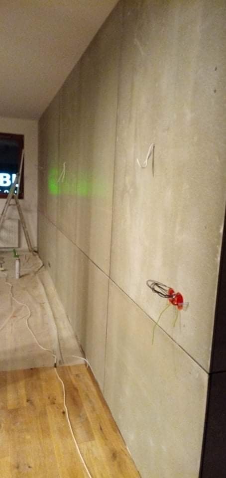 Montaż płyt karton gips na ścianie, widoczne kable elektryczne wystające ze ściany, drabina oparta o ścianę przy oknie, podłoga drewniana.