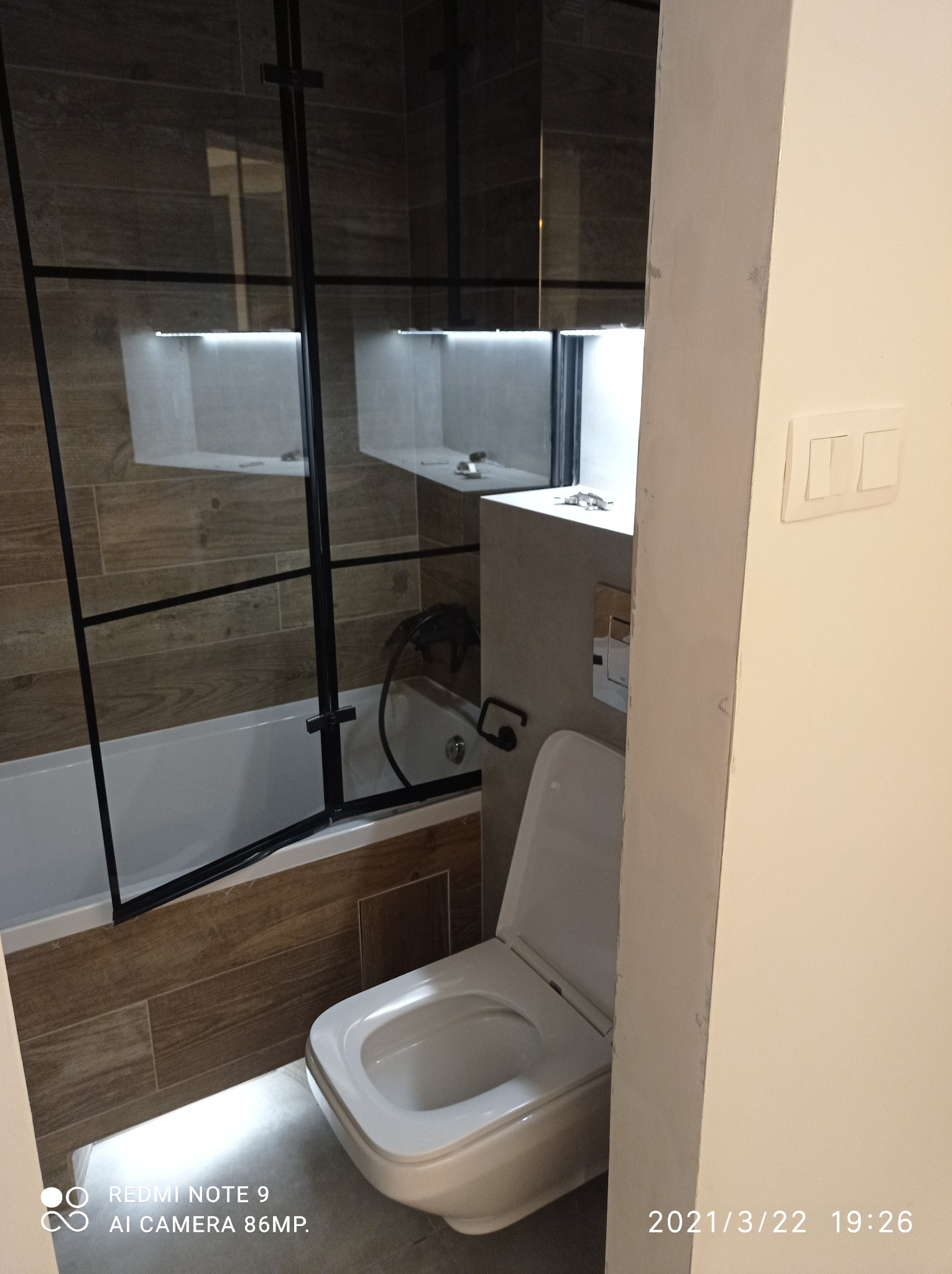 Wykończona łazienka z kabiną prysznicową typu walk-in z czarnymi ramami, toaletą i płytkami imitującymi drewno na ścianach.