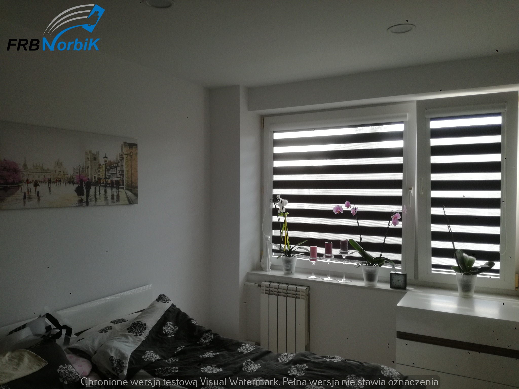 Sypialnia z łóżkiem, grzejnikiem pod oknem z roletami dzień-noc, dekoracjami na parapecie i obrazem na ścianie, oświetlona światłem dziennym.