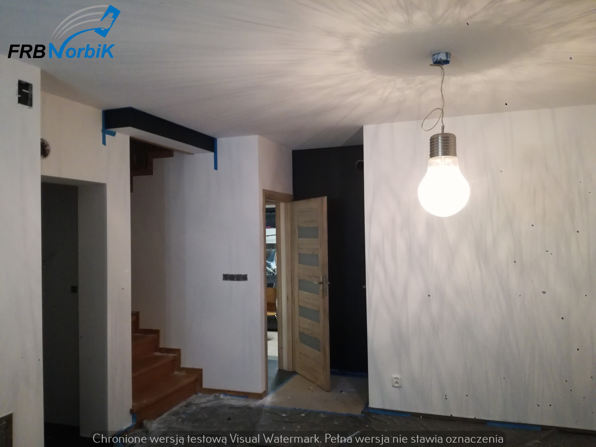 Wnętrze w trakcie odświeżania: białe ściany z widocznymi niedoskonałościami, schody zabezpieczone folią, surowa żarówka wisząca na kablu, logo firmy w rogu.