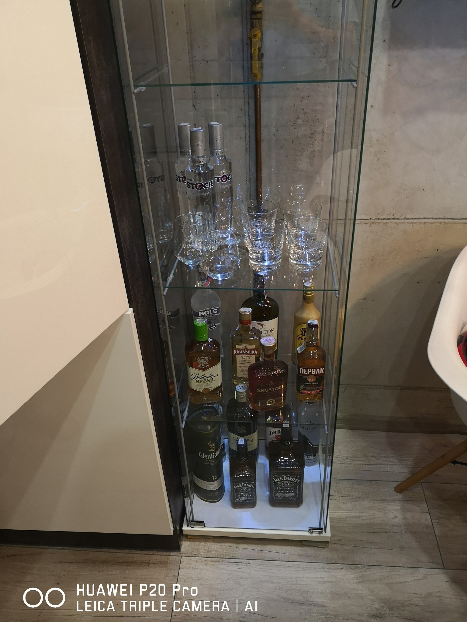 Szklana witryna z oświetleniem eksponująca kolekcję alkoholi, w tym butelki Jack Daniel's i Ballantine's, oraz zestaw kryształowych szklanek, z widoczną instalacją hydrauliczną w tle.