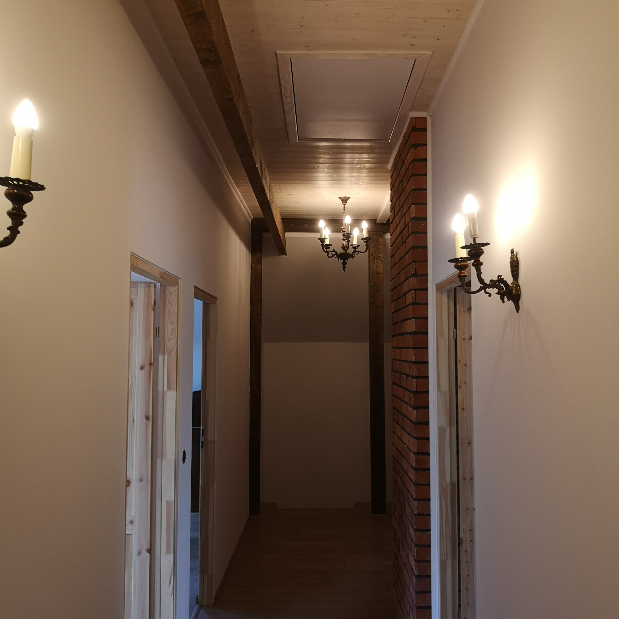Korytarz z drewnianym sufitem podwieszanym, ceglaną ścianą, drewnianymi belkami i ozdobnymi kinkietami w stylu retro.