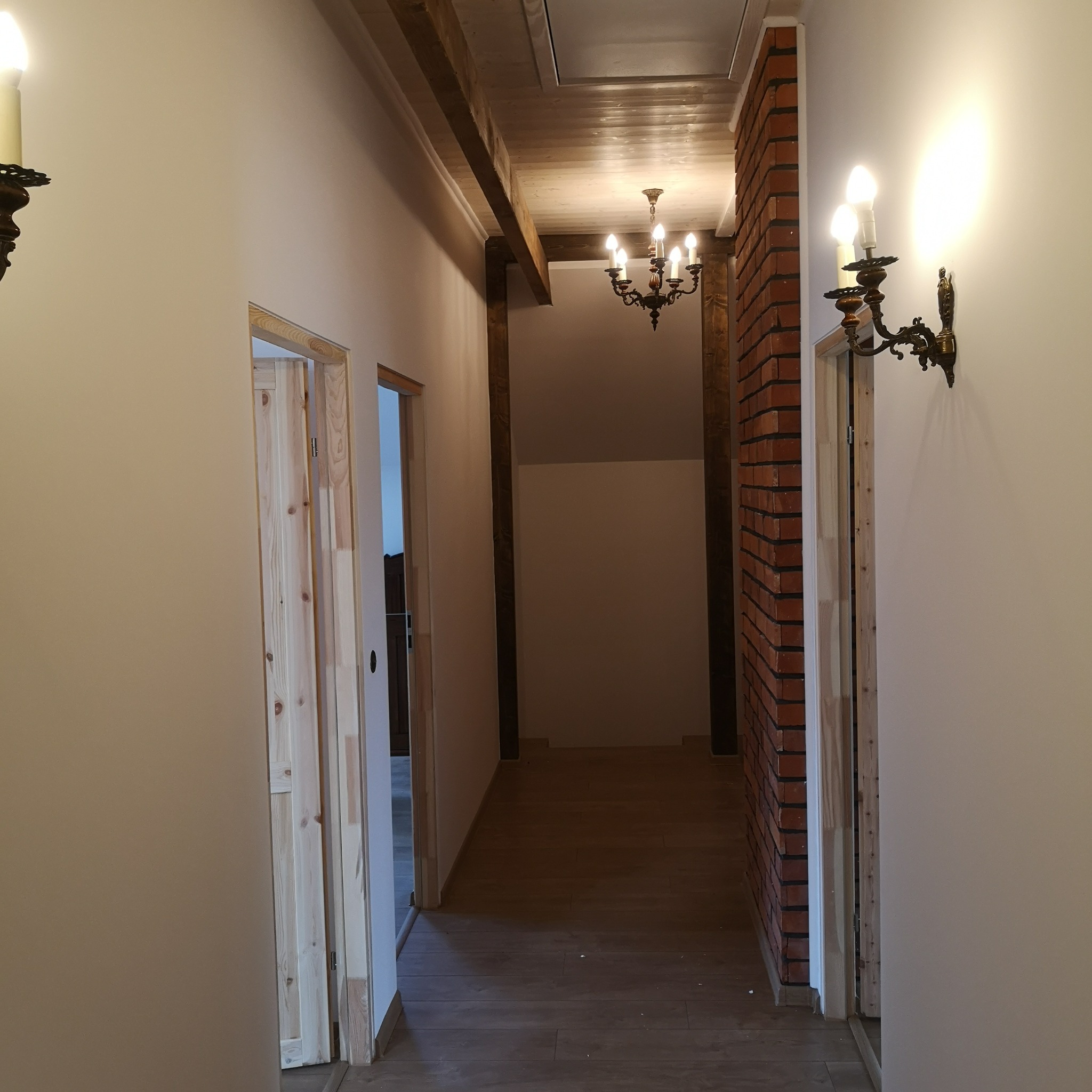 Korytarz w domu z drewnianymi belkami stropowymi, ceglaną ścianą i klasycznymi kinkietami, widoczne drzwi w trakcie montażu ościeżnic.