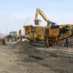 Żółta kruszarka do gruzu EXTEC z koparką CAT załadowuje materiał, w tle dwie inne maszyny budowlane na placu budowy.