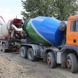 Dwa samochody betonomieszarki na placu budowy, jeden biały z wysięgnikiem, drugi pomarańczowy z bębnem w kolorach niebieskim i zielonym, na tle drzew i bloku mieszkalnego.