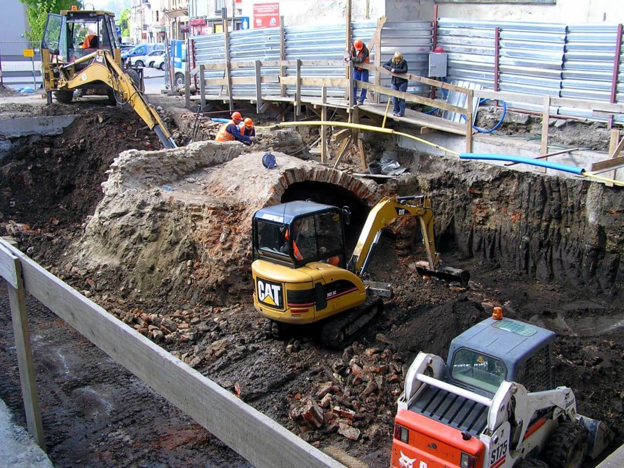 Prace ziemne przy odsłanianiu starego, ceglanego łuku. Na budowie obecne koparki marki CAT i Bobcat oraz pracownicy w kaskach.