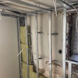 Wnętrze w trakcie prac: metalowy stelaż pod płytę karton-gips, częściowo wypełniony wełną mineralną, widoczne kable elektryczne i fragment starej, ceglanej ściany.
