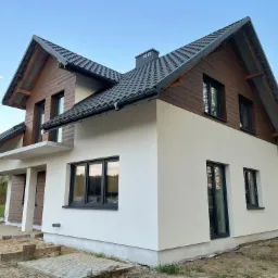 Dwukondygnacyjny dom z wykończoną elewacją: biały tynk na dole i brązowe panele imitujące drewno na górze, czarne okna i dachówka, widoczne prace brukarskie wokół budynku.
