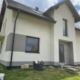 Nowoczesny dom jednorodzinny z dwukolorową elewacją, widok z boku na świeżo pomalowaną fasadę, z widocznymi materiałami budowlanymi na trawniku i elementami wykończeniowymi.