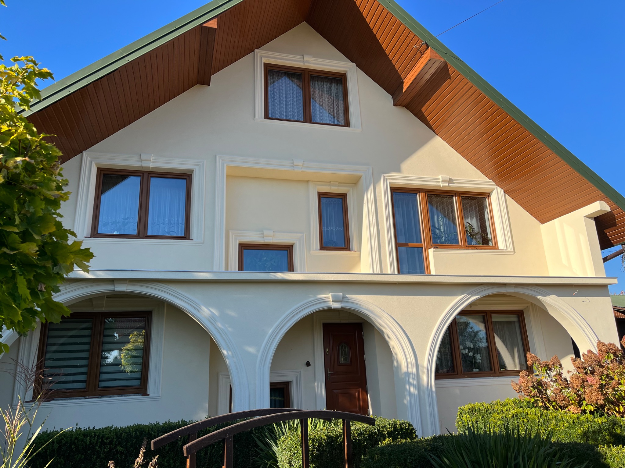 Dwupiętrowy dom z jasną elewacją, brązową stolarką okienną i zielonym dachem, widoczny z poziomu ogrodu z ozdobnymi krzewami i metalowym mostkiem.