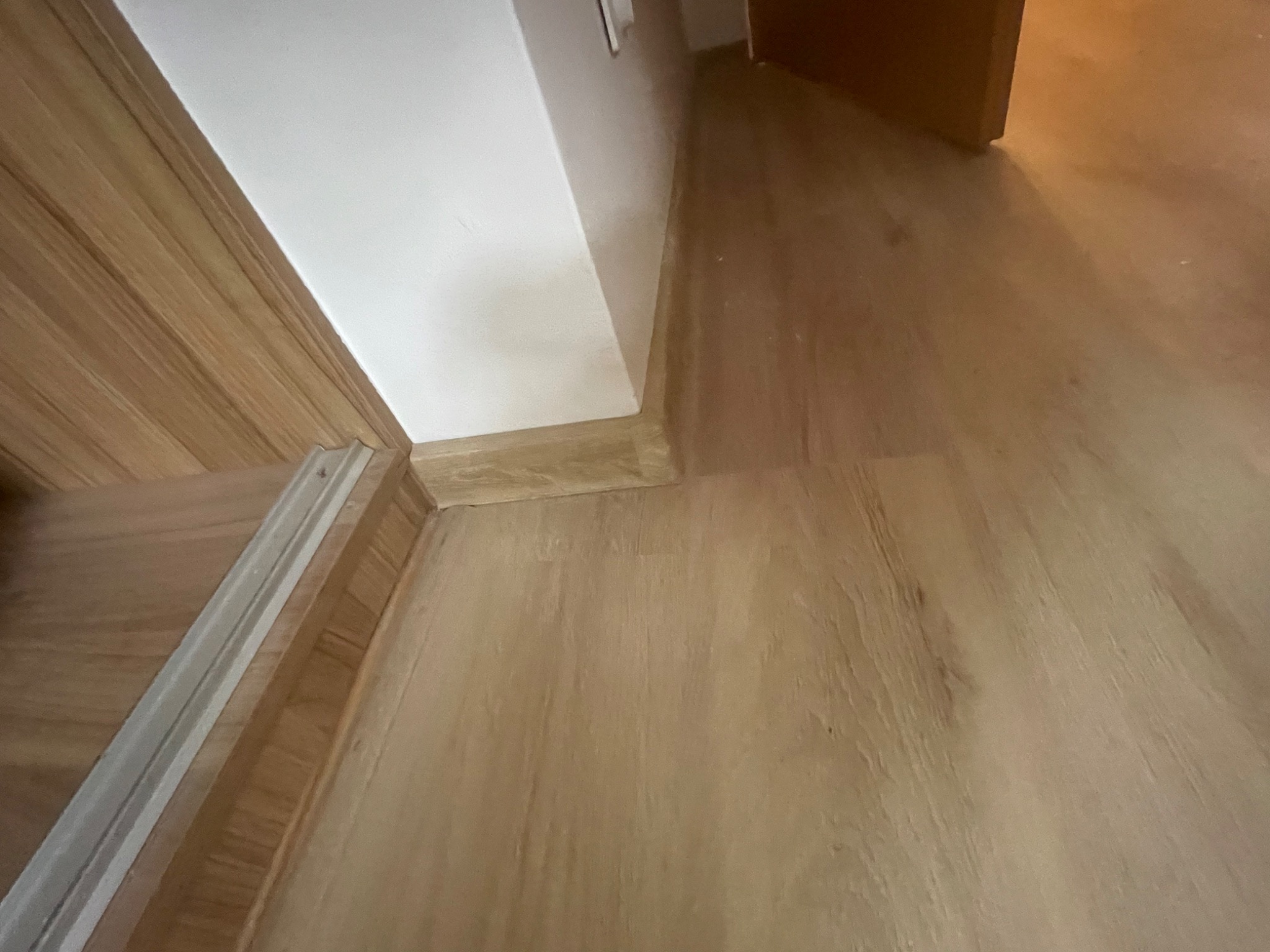 Fragment wnętrza z drewnopodobną podłogą, białą ścianą i listwą przypodłogową, widok z niskiej perspektywy.