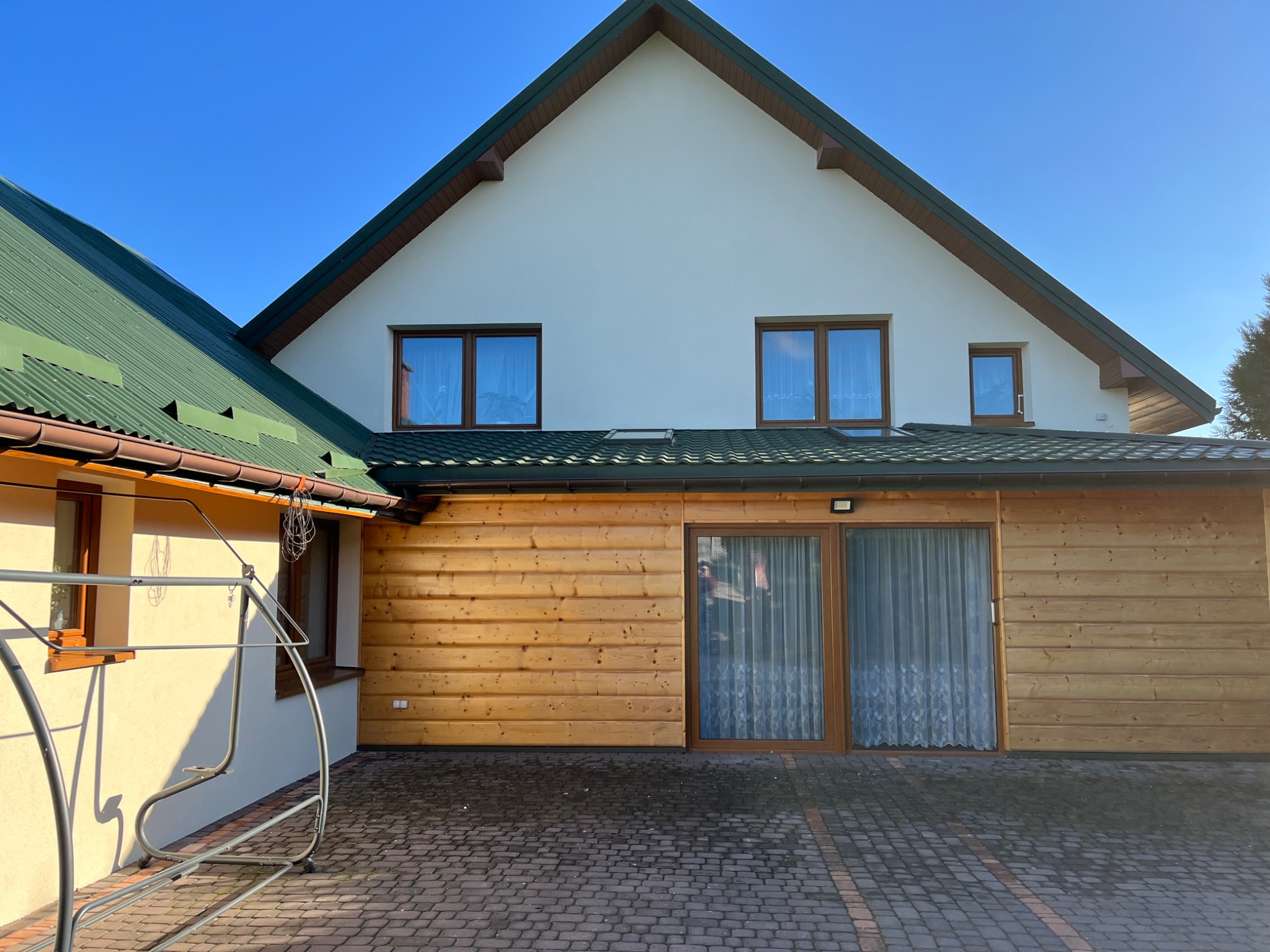 Dwukondygnacyjny dom z jasną elewacją i zielonym dachem, część ściany wykończona drewnianą boazerią, widoczne okna z brązowymi ramami, na pierwszym planie fragment huśtawki.