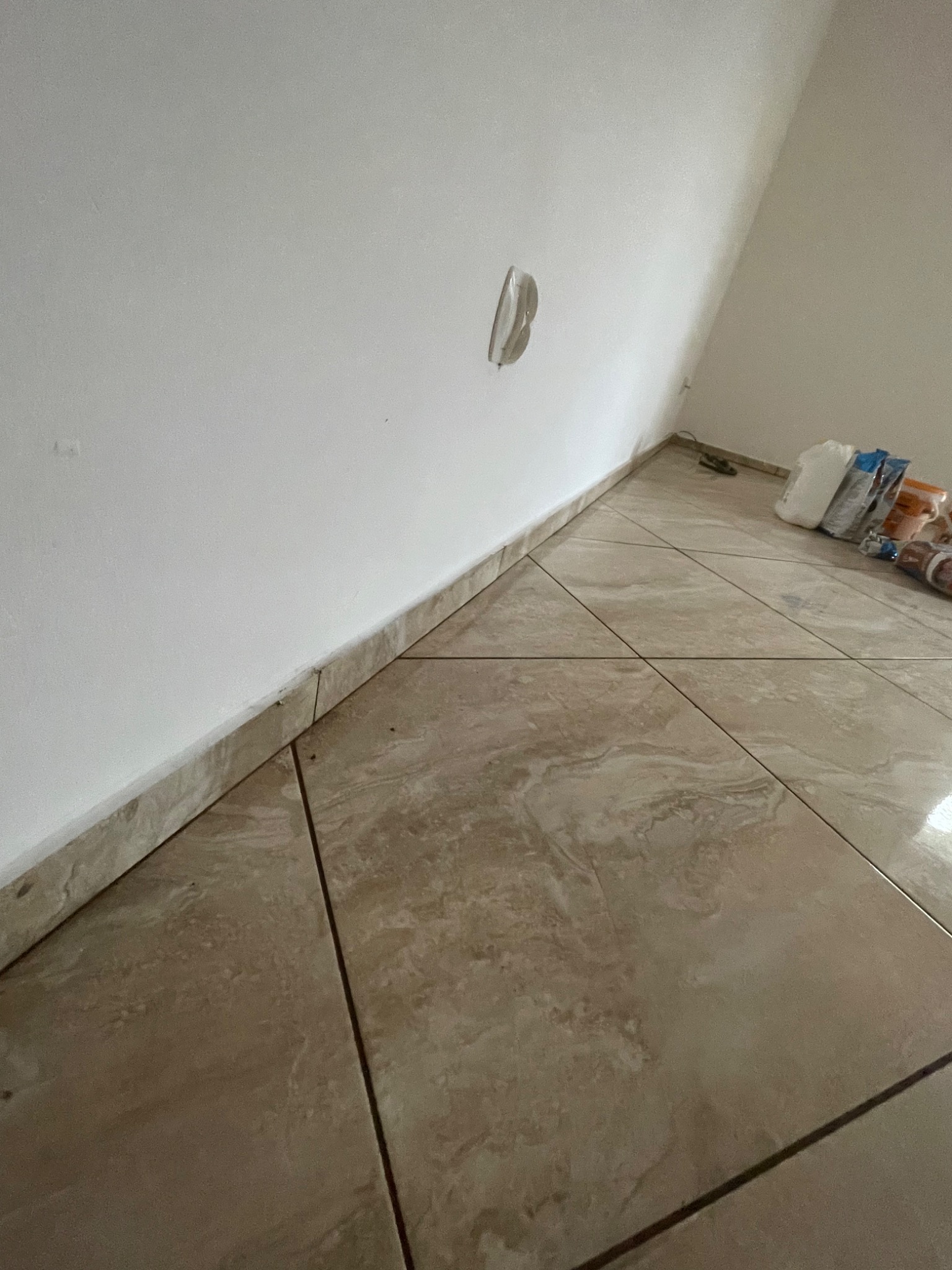 Świeżo pomalowana biała ściana z wystającym kablem elektrycznym i ułożona beżowa podłoga z płytek ceramicznych. W tle widoczne materiały budowlane.