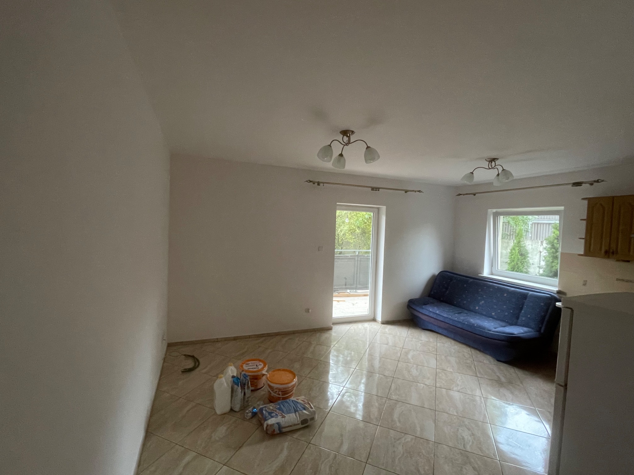 Wnętrze pokoju przed malowaniem: widoczne przygotowane farby, grunt i akcesoria malarskie na podłodze z płytek, pusta ściana, niebieska sofa pod oknem, sufit z lampami.