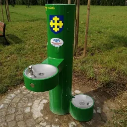 Zielony, metalowy źródełko wody pitnej z dwoma misami (dla ludzi i zwierząt) na tle trawnika i drzew. Widoczne herby i napisy na obudowie.