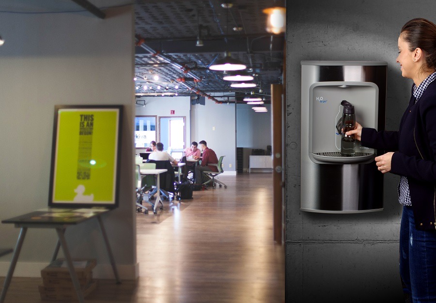 Kobieta napełnia bidon z dystrybutora wody w nowoczesnym biurze z widocznym open space w tle.