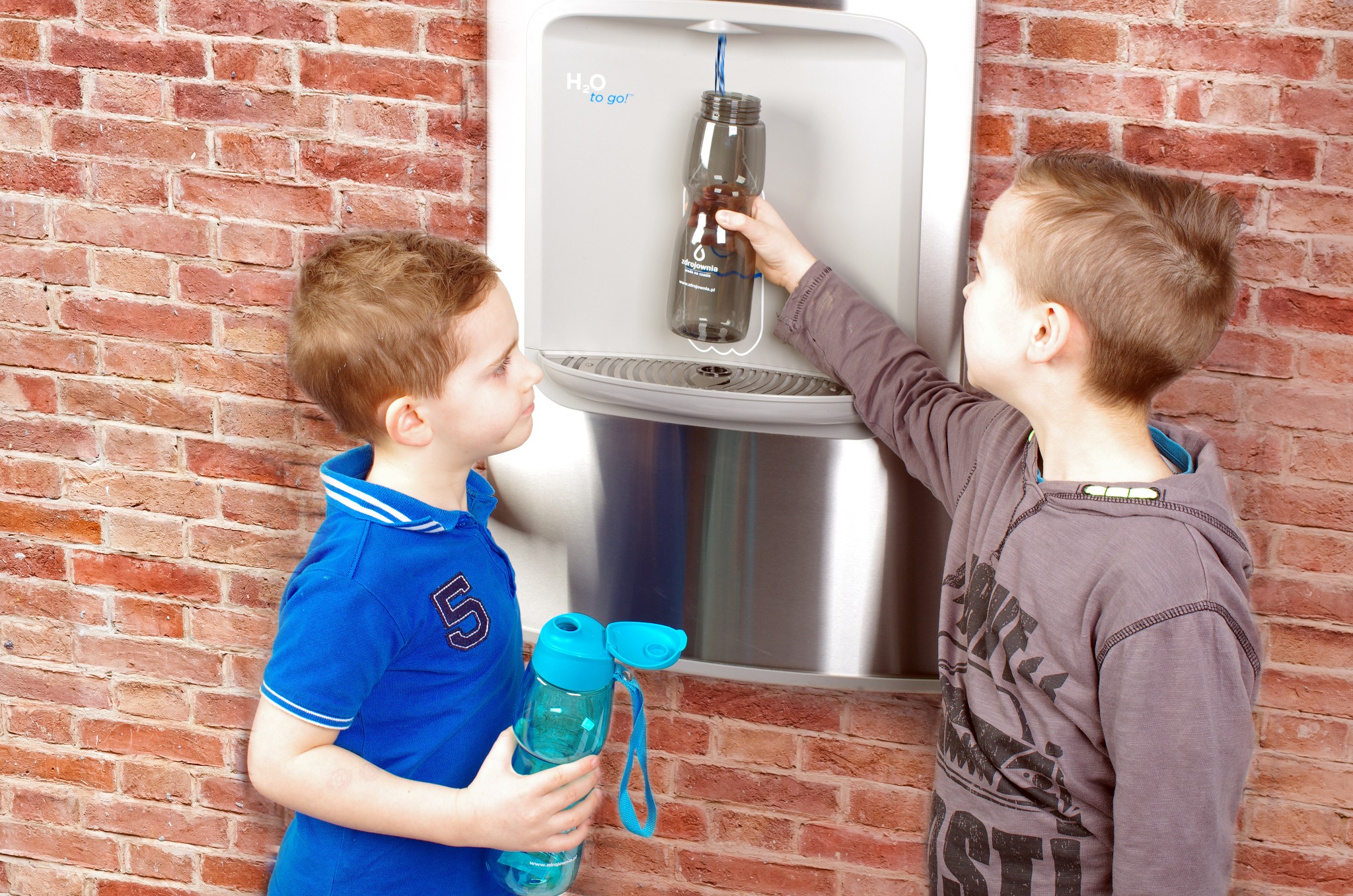 Dwóch chłopców przy dystrybutorze wody H2O to go!, jeden napełnia butelkę, drugi obserwuje, na tle ceglanej ściany.