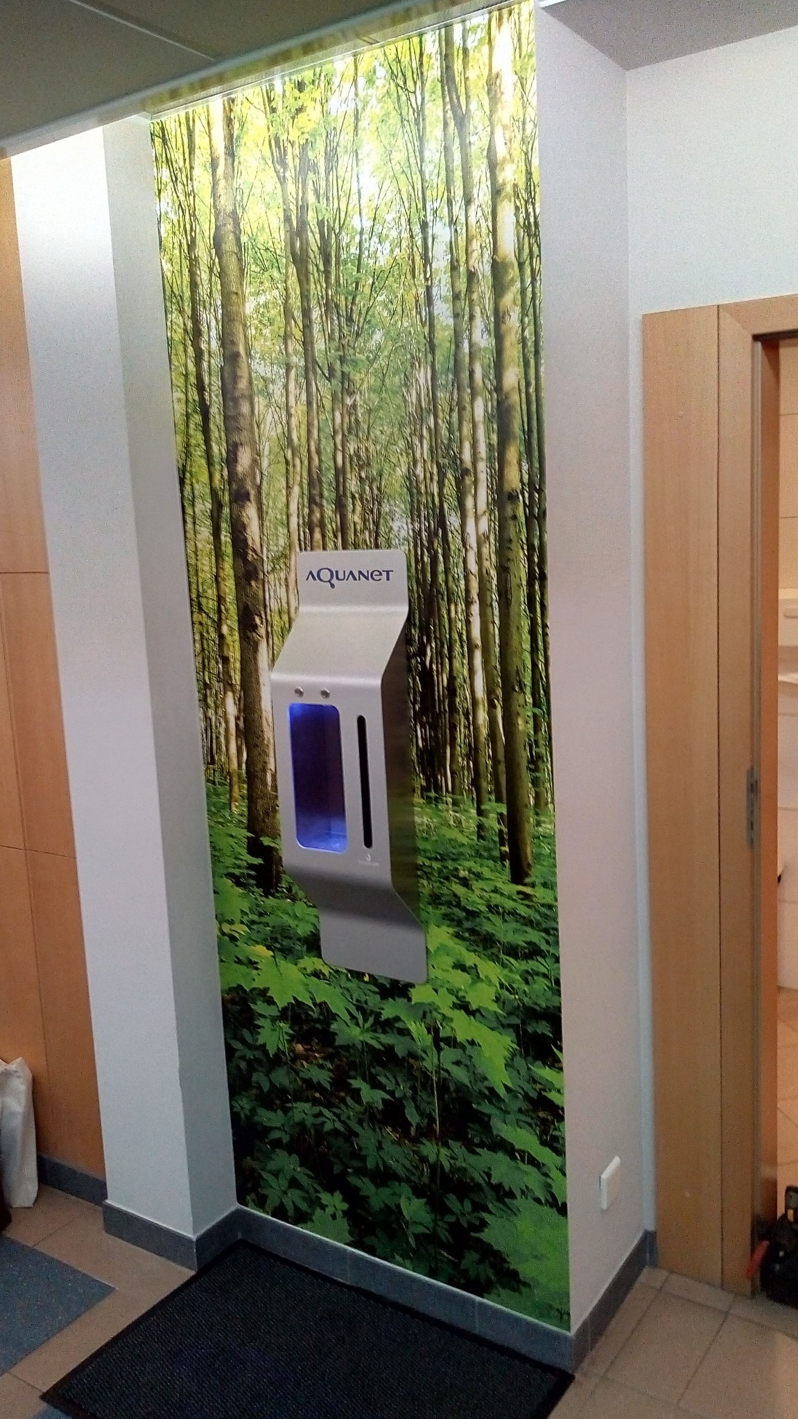 Dozownik wody Aquanet na ścianie z fototapetą leśną, widoczny na tle zieleni drzew, z niebieskim podświetleniem wewnątrz.