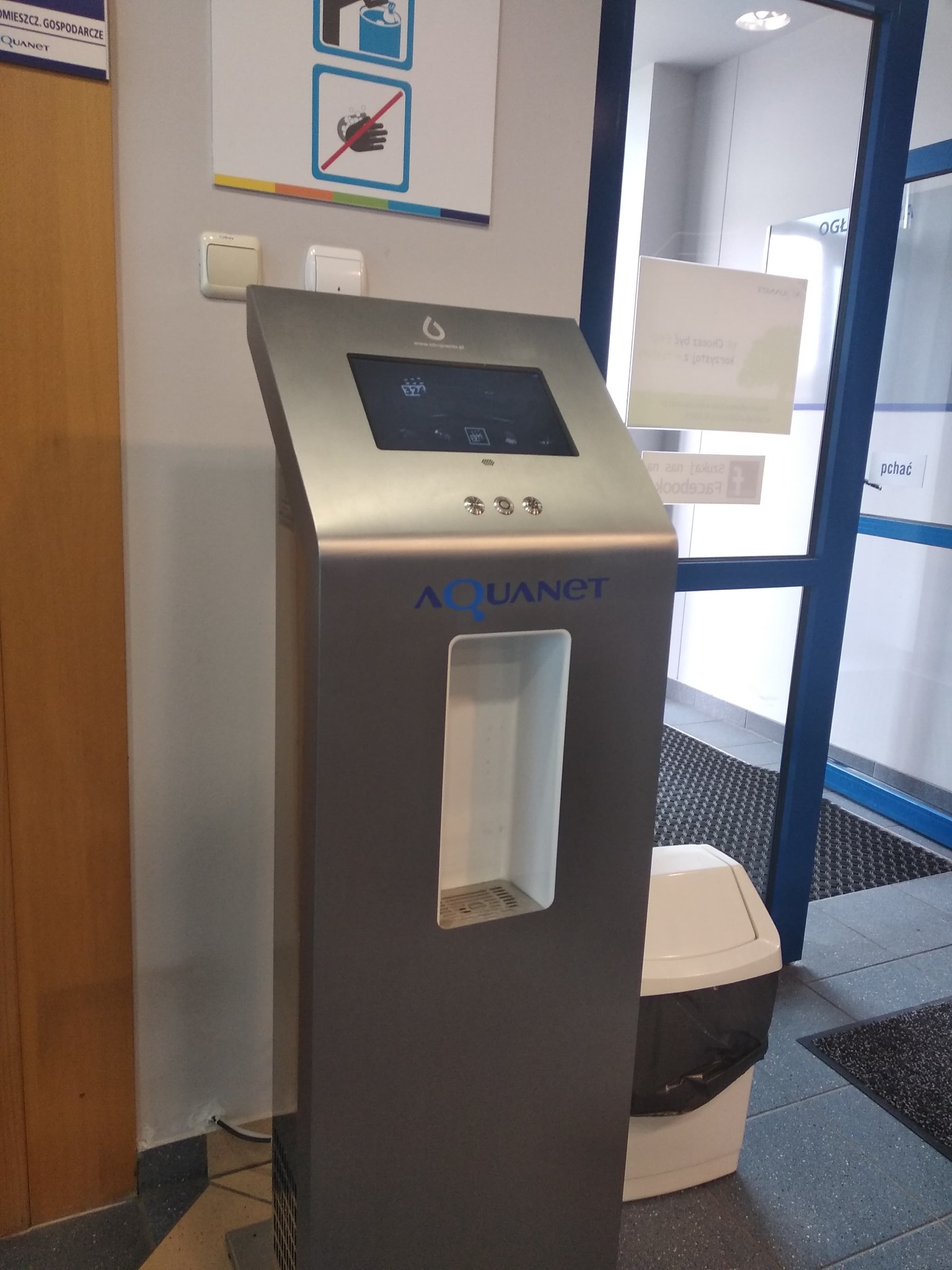Nowoczesny dystrybutor wody Aquanet z ekranem dotykowym, umieszczony w przestrzeni biurowej, obok kosza na śmieci i niebieskich drzwi wejściowych.