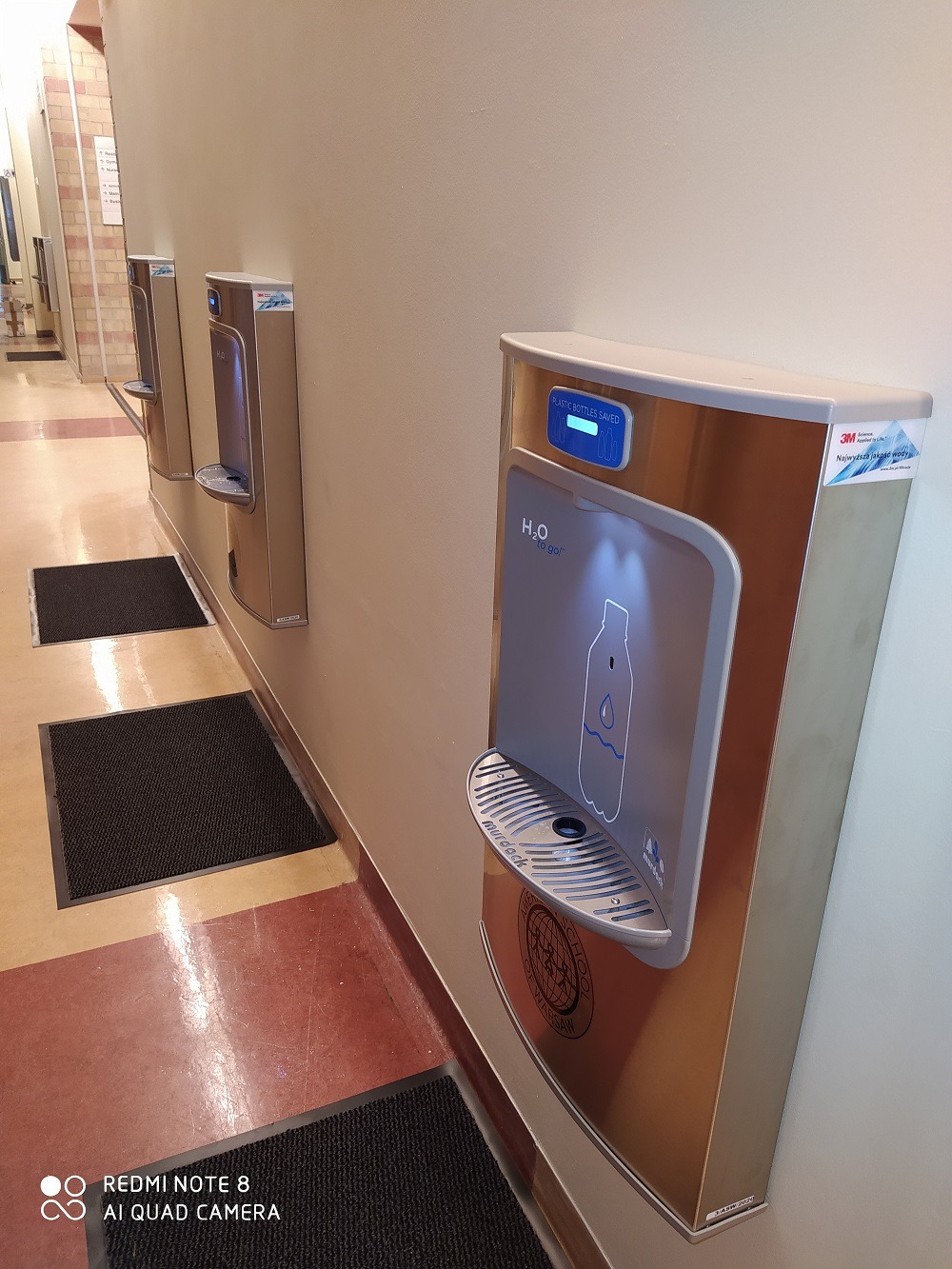 Bezkontaktowe dystrybutory wody pitnej marki Murdock zamontowane na ścianie w korytarzu, z wyświetlaczem ilości zaoszczędzonych plastikowych butelek.