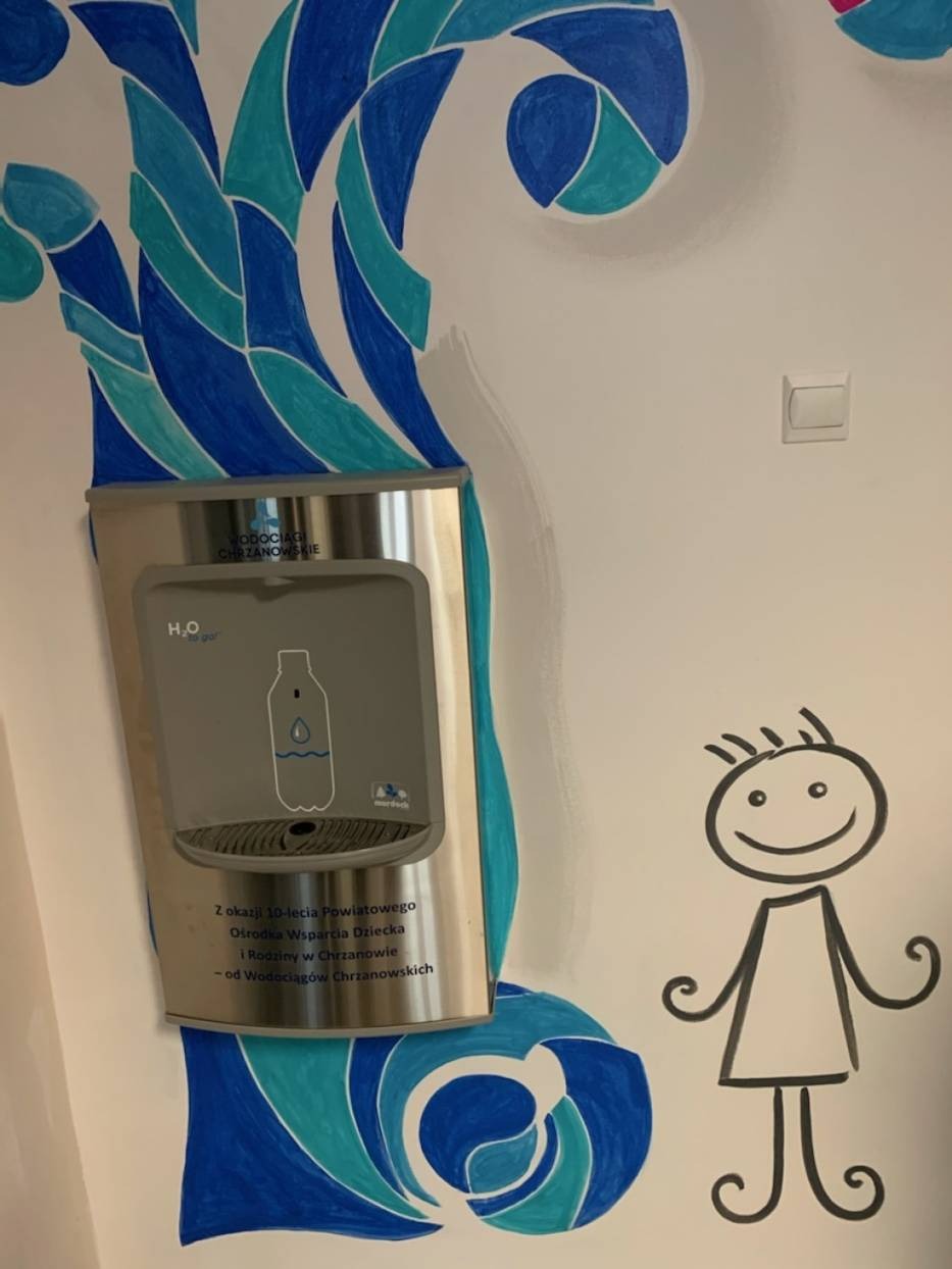 Dystrybutor wody pitnej z napisem 'Wodociągi Chrzanowskie' umieszczony na ścianie z rysunkiem stylizowanego drzewa i postaci dziecka.
