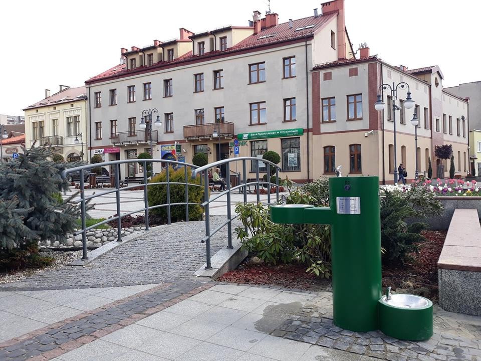 Zewnętrzny, publiczny, zielony automat z pitną wodą na tle miejskiego placu z budynkami i zielenią.