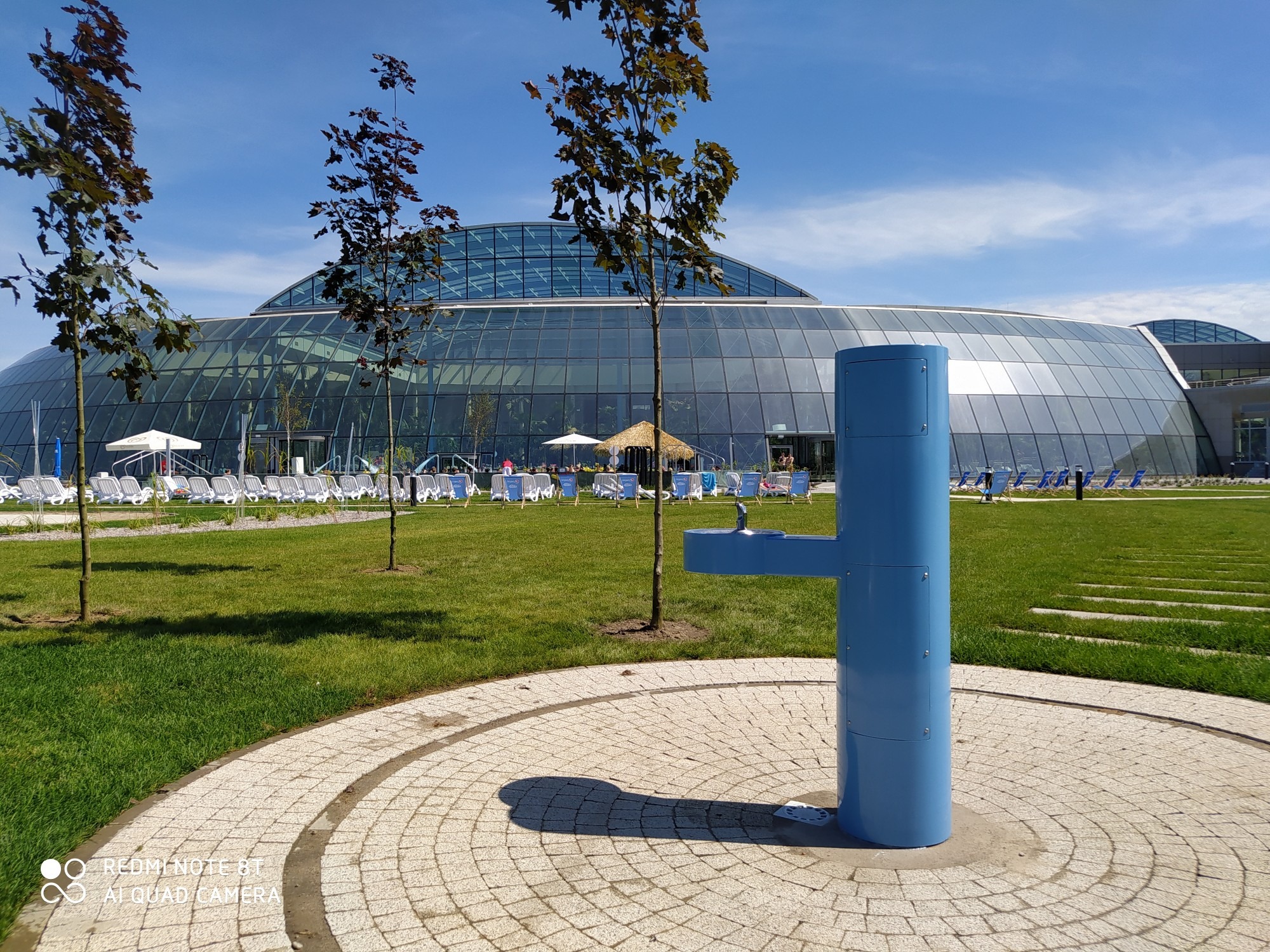 Niebieski dystrybutor wody pitnej na tle nowoczesnego aquaparku z przeszkloną kopułą, otoczonego zielonym trawnikiem i leżakami.