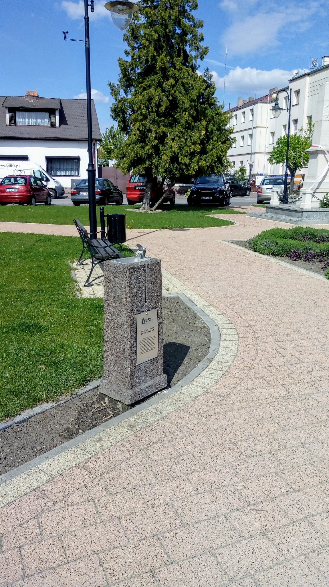 Zewnętrzny, kamienny dystrybutor wody pitnej z metalowym kranikiem w przestrzeni publicznej, na tle trawnika, drzewa i parkingu.