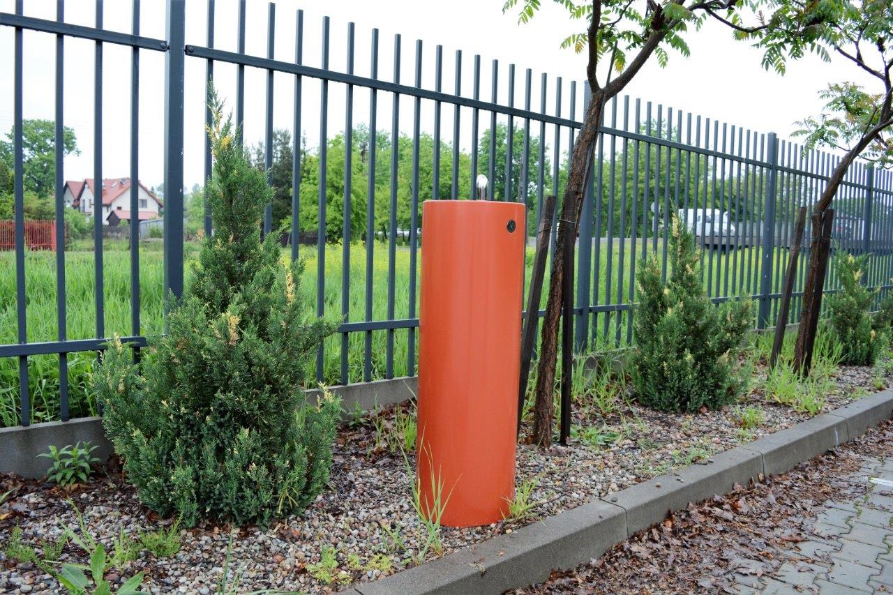 Pomarańczowy hydrant zewnętrzny na terenie posesji w Zielonce, otoczony zielenią i ogrodzeniem.