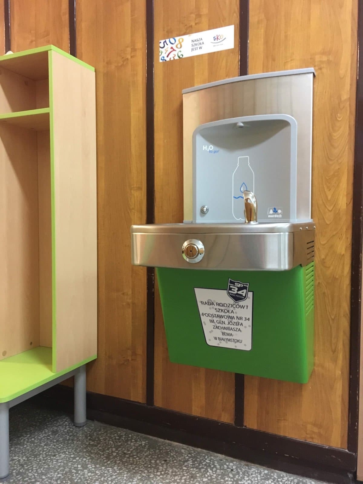 Stalowa fontanna wody pitnej z zielonym pojemnikiem na ścianie w szkole, obok szafka z jasnego drewna z zielonymi elementami.