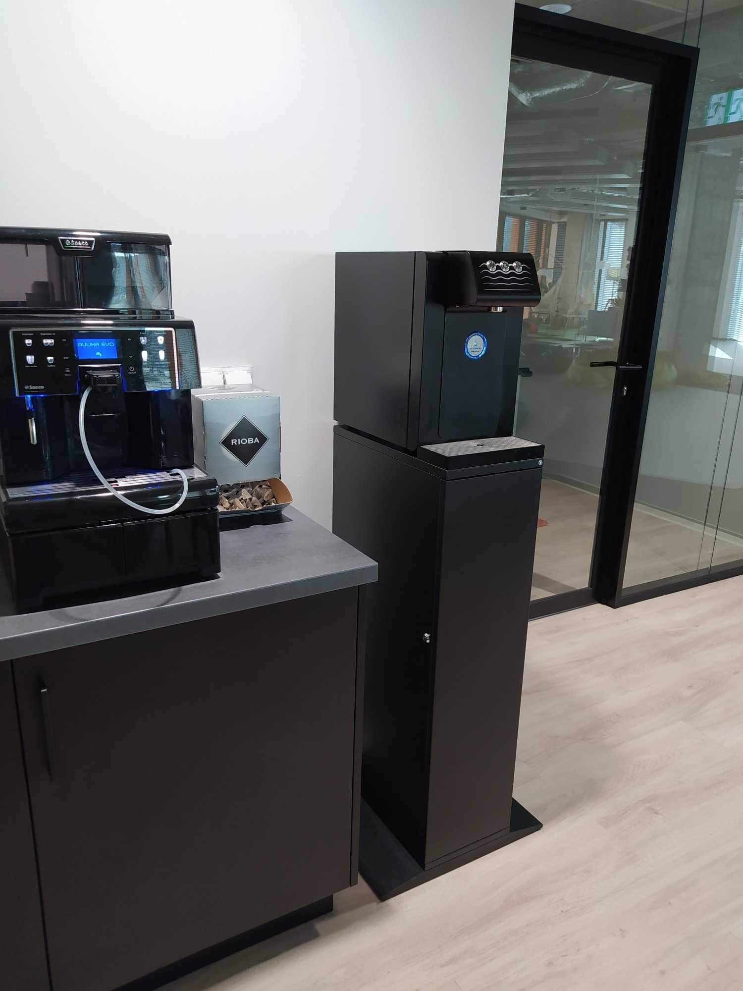 Automatyczny dystrybutor wody butelkowanej z ekspresem do kawy Saeco Aulika Evo, ustawiony w biurze obok szklanej ściany działowej.