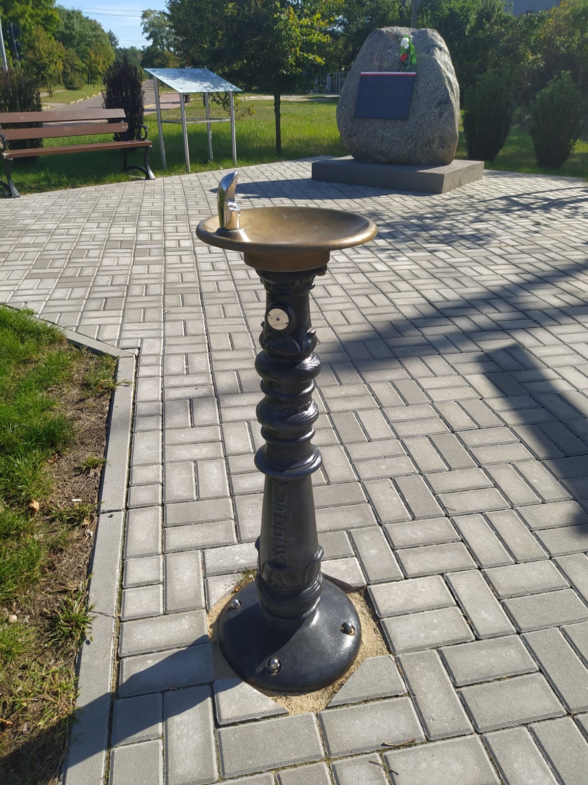 Zdrój uliczny z metalową misą i ozdobną, żeliwną kolumną na tle parku z pomnikiem i ławkami.