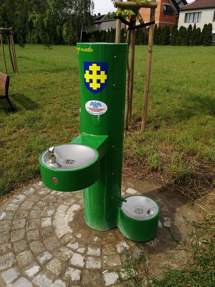 Zielony, metalowy źródełko wody pitnej z dwoma misami (dla ludzi i zwierząt) na tle trawnika i drzew. Widoczne herby i napisy na obudowie.