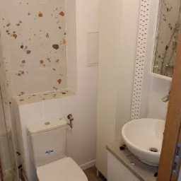 Wykończona łazienka z toaletą, umywalką nablatową i płytkami imitującymi lastryko na ścianie, widoczne białe meble łazienkowe i dekoracyjny pasek na ścianie.