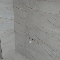 Wyłożona beżowymi płytkami imitującymi marmur ściana i podłoga w rogu łazienki, widoczne otwory na instalacje wodne i elektryczne.