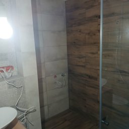 Snowbud Bartosz Śnieżewski - Wykończona łazienka z kabiną prysznicową i płytkami imitującymi drewno. Widoczne przyłącza wodne i umywalka z szafką. Oświetlenie górne.