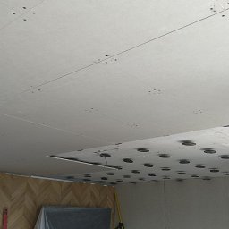 Snowbud Bartosz Śnieżewski - Sufit z płyt kartonowo-gipsowych z widocznymi otworami na oświetlenie, ściana z wzorem jodełki częściowo zakryta folią ochronną, narzędzia w rogu.