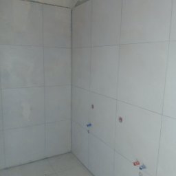 Snowbud Bartosz Śnieżewski - Wykończona, jasna łazienka z białymi, kwadratowymi płytkami na ścianach i podłodze. Widoczne otwory na instalacje wodne. Surowy stan wykończenia sufitu.